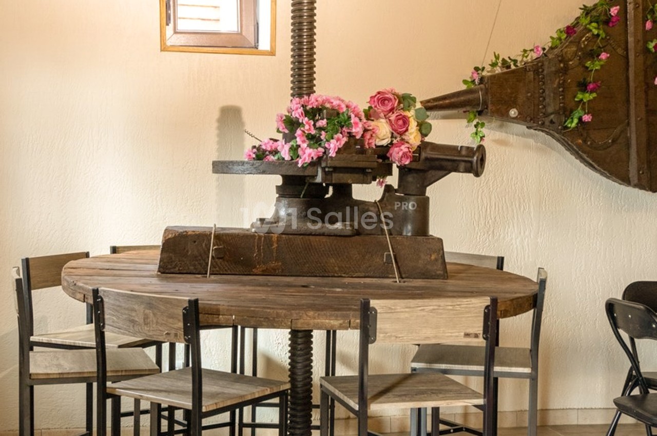 Table ronde en bois entourée de chaises en bois et métal, avec un ancien pressoir en fer fleuri au centre.