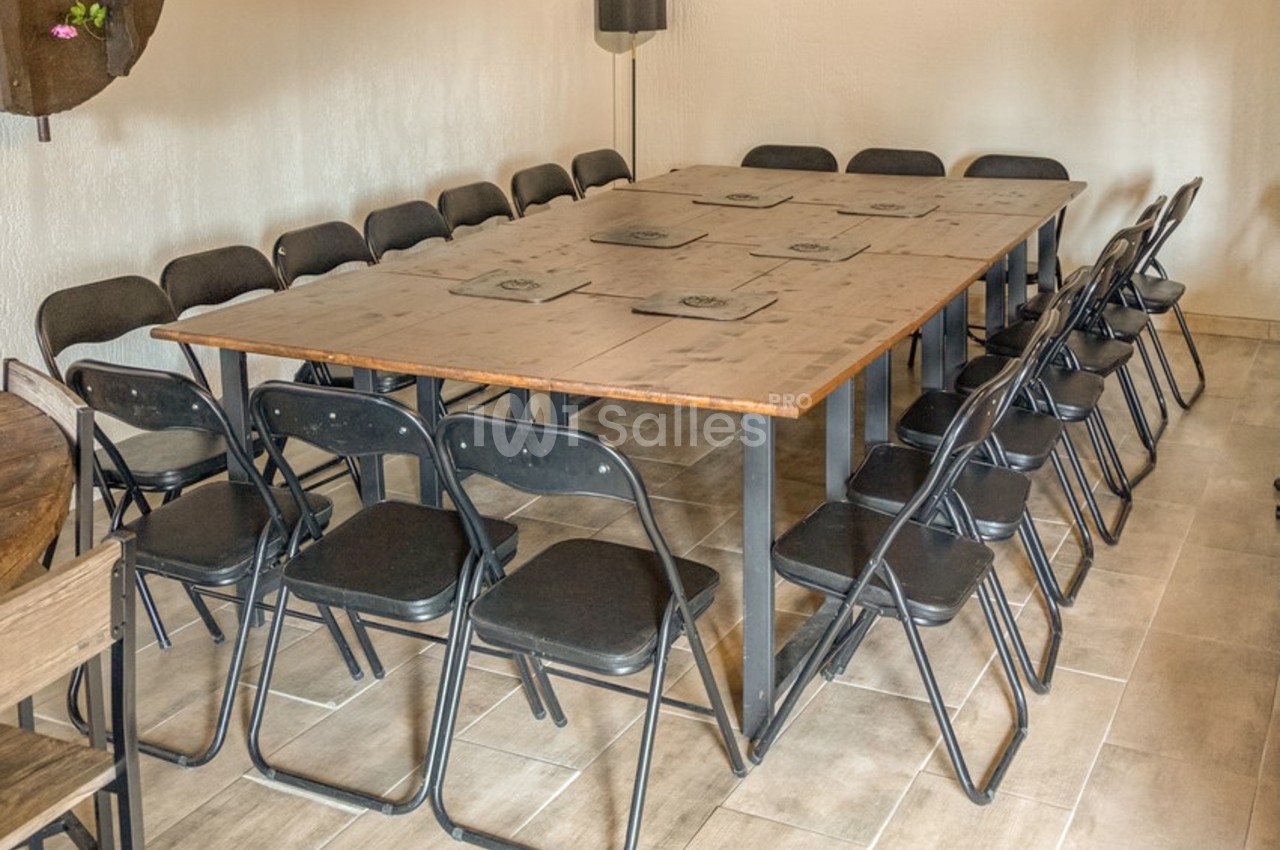 Salle aux murs clairs avec une grande table rectangulaire en bois entourée d'une vingtaine de chaises pliantes noires, sol carrelé. Let me check length: roughly 130 characters? Count: "Salle aux murs clairs avec une grande table rectangulaire en bois