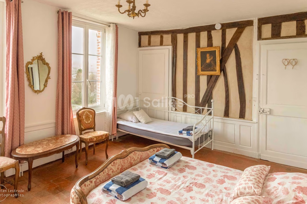 Chambre ancienne aux murs à colombages, avec un grand lit recouvert d'un tissu à coquillages roses, un petit lit de fer blanc contre le mur et un mobilier de style Louis XV. Let me check length: roughly 165 characters, too long. Need ≤125. Chambre