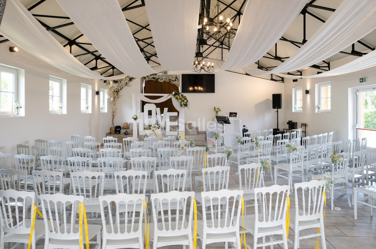 Salle de réception lumineuse et blanche, dressée pour une cérémonie avec rangées de chaises blanches à rubans jaunes, plafond drapé de voilages et estrade fleurie au fond.
