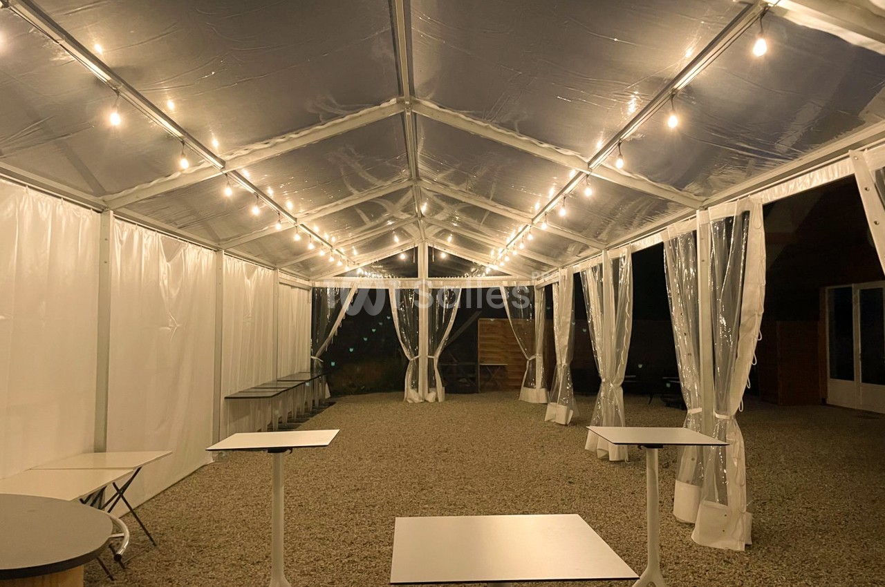 Espace sous chapiteau transparent la nuit, éclairé de guirlandes lumineuses, avec tables hautes blanches, parois et rideaux translucides sur un sol en gravier. Let me check length: roughly 165 characters - too long. Need ≤125. Shorter: "Chapiteau