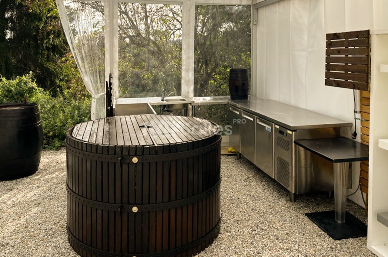 Tente au sol de gravier abritant un grand tonneau en bois, un plan de travail inox et des réfrigérateurs professionnels.