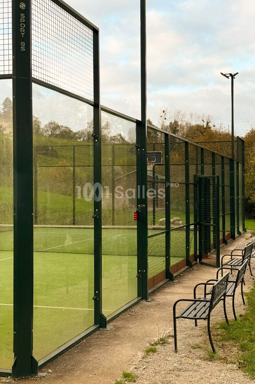 Court de padel extérieur à surface verte, entouré de parois vitrées et grillagées sombres, avec un banc en métal le long du cheminement de gravier.