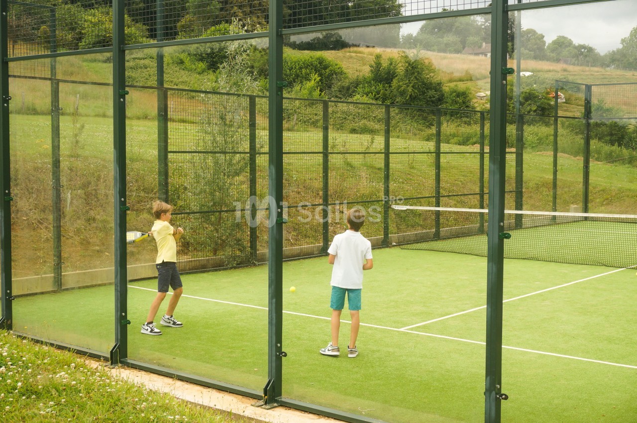 Deux enfants jouent au padel sur un court vert entouré de grillages et de vitres, avec une prairie et des arbres en arrière-plan.