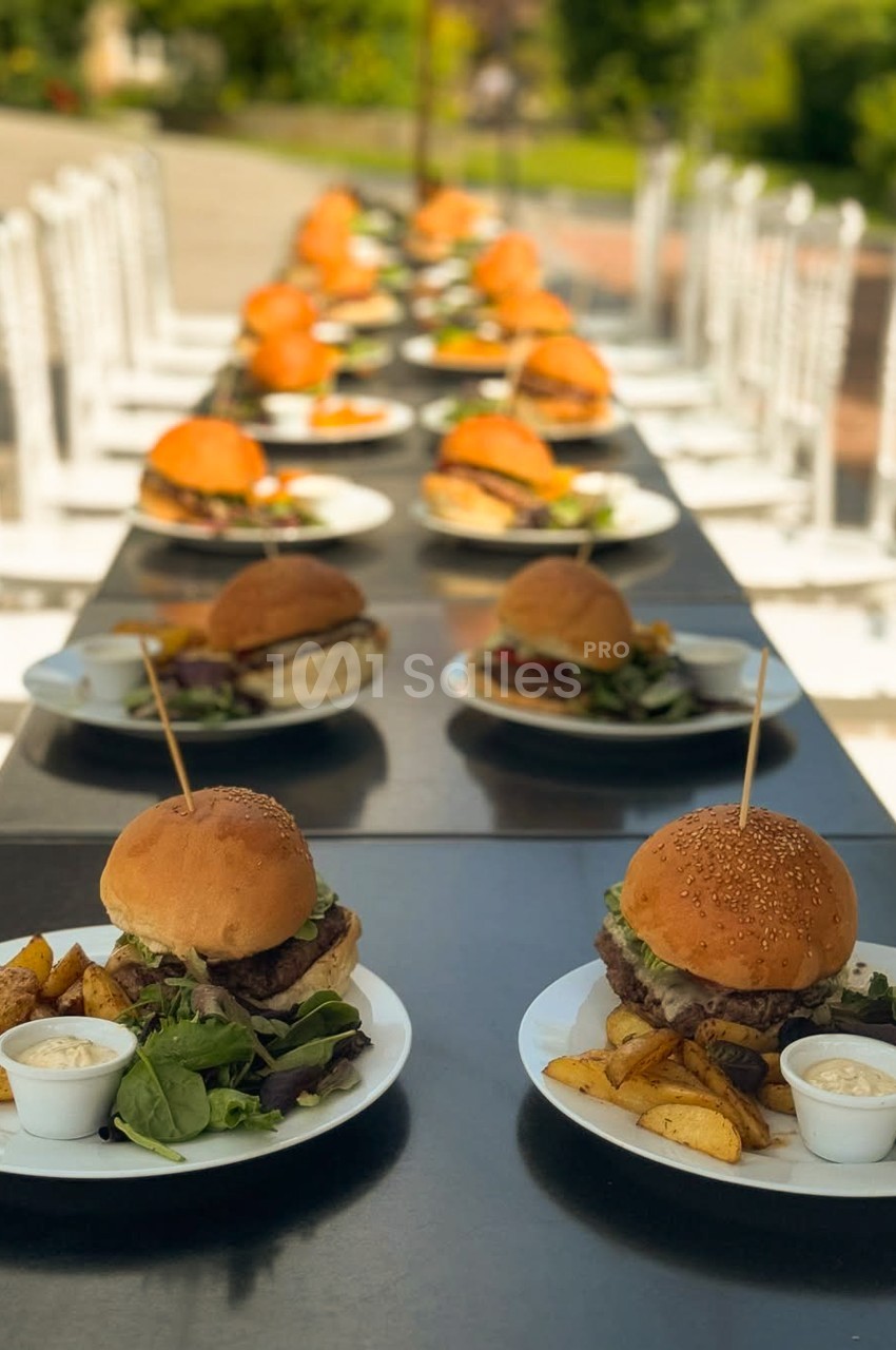 Burgers garnis de frites et de salade, disposés en rang sur une longue table extérieure, avec des chaises blanches en arrière-plan.