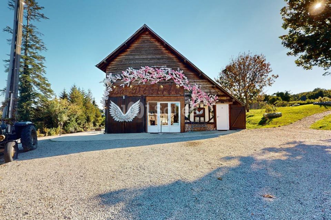 Bâtiment en bois à toit à deux pans, orné de fleurs roses artificielles et d'une grande aile blanche peinte sur une porte, sur une aire de gravier. Let me check: under 125 chars? Count roughly: "Bâtiment en bois à toit à deux pans, orné de fleurs roses