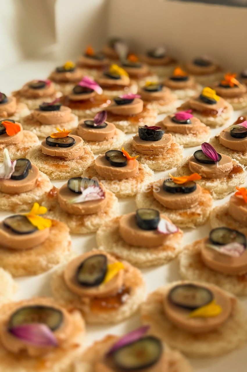 Rangées de canapés sur toasts ronds, garnis d'une tranche de pâté, d'une rondelle sombre et de fleurs comestibles.