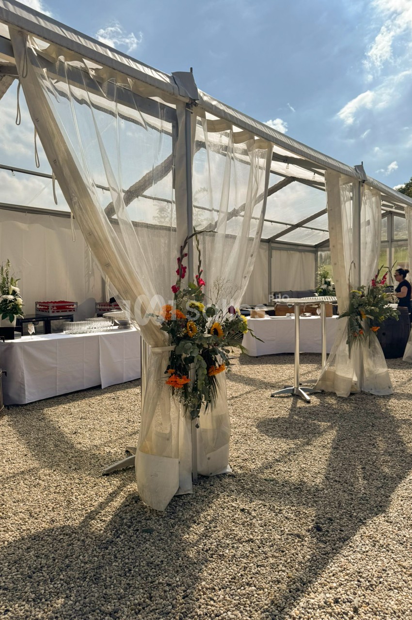Structure de réception sous tente transparente, drapés de voilage ivoire ornés de bouquets de fleurs orange et jaunes, tables de buffet nappées en arrière-plan.