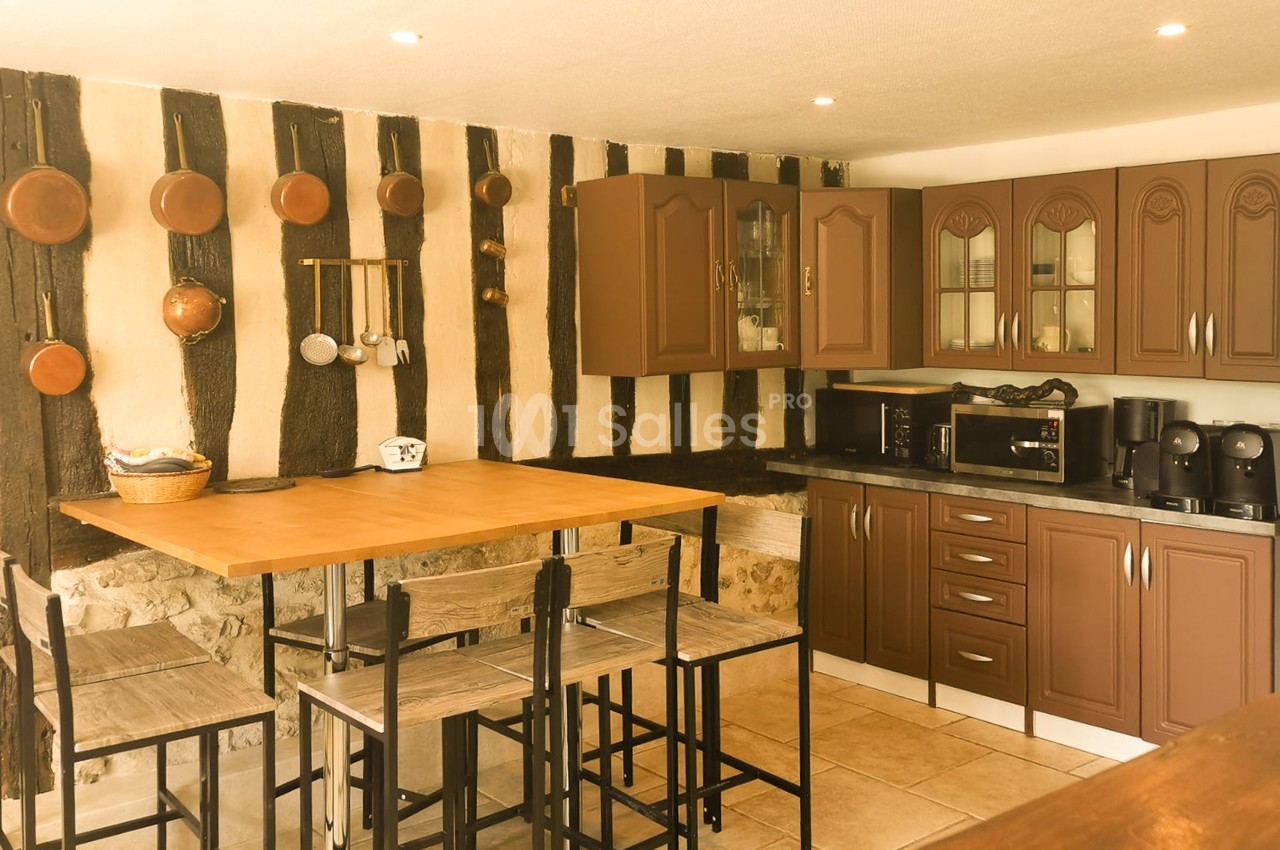 Cuisine rustique avec casseroles en cuivre accrochées à un mur à colombages, table haute en bois entourée de tabourets, et meubles de cuisine marron à côté.