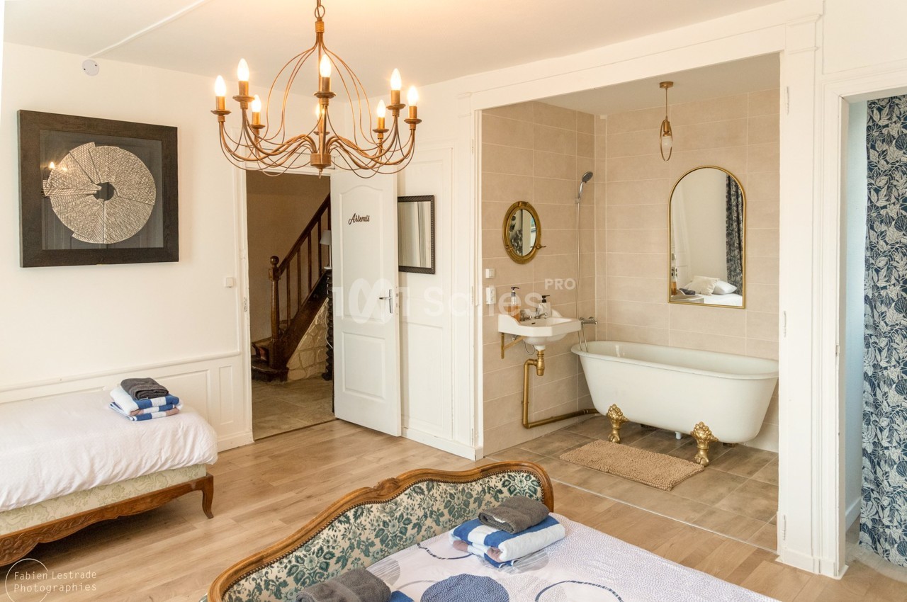 Chambre lumineuse avec lustre doré, lit et méridienne, ouverte sur une salle de bain à baignoire sur pieds.