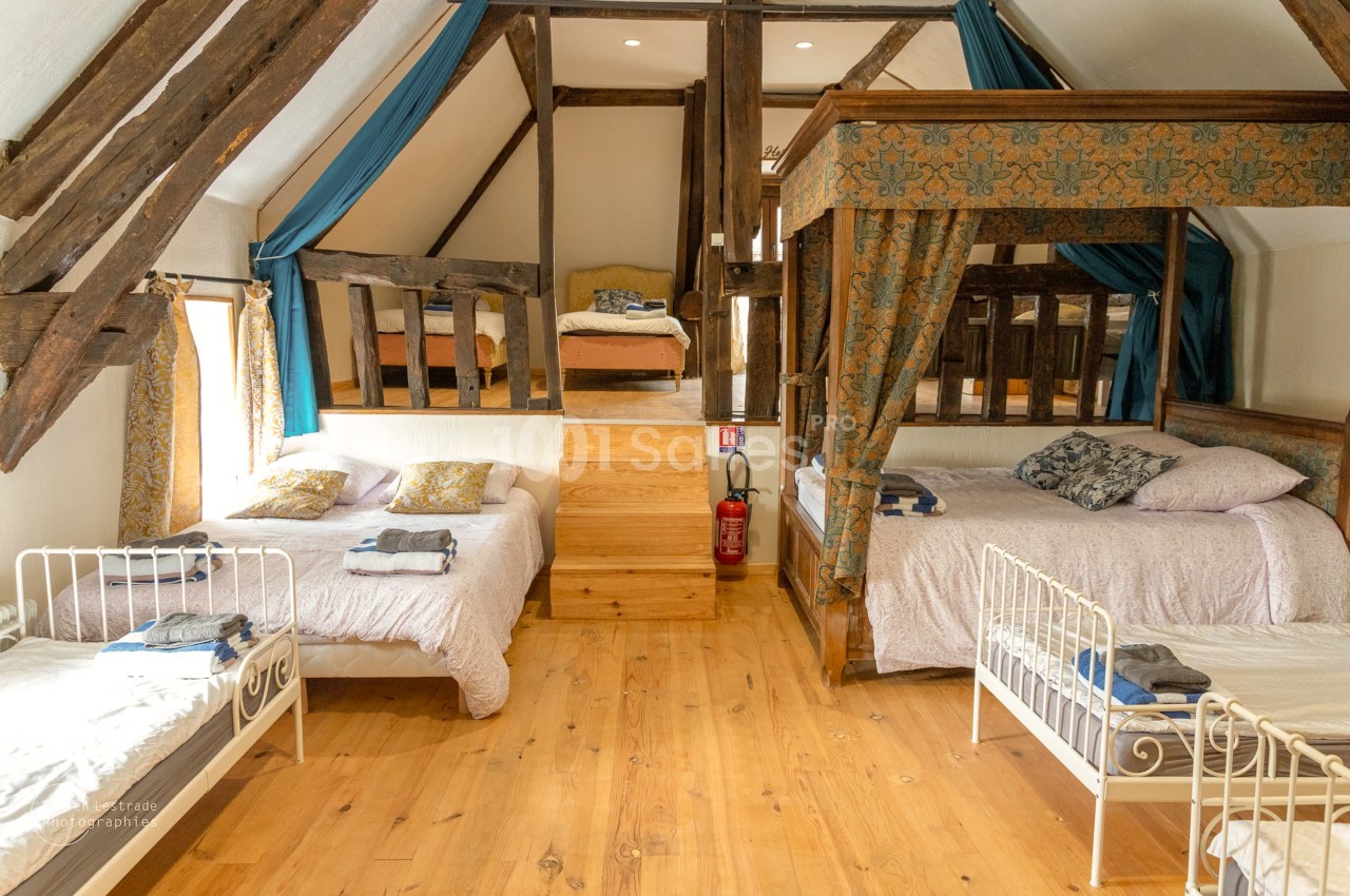 Chambre mansardée aux poutres apparentes avec plusieurs lits : un lit à baldaquin en bois, deux lits doubles et des lits simples en fer forgé.