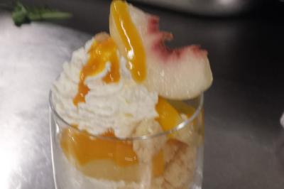 Verrine de dessert composée de crème fouettée, morceaux de pêche et coulis de mangue sur un lit de biscuit.