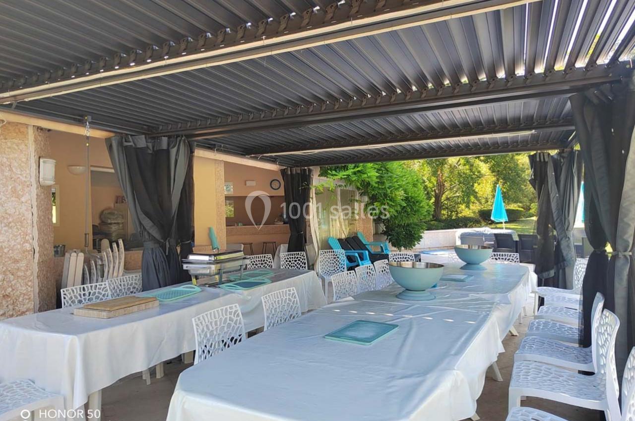 Tables dressées avec nappes blanches sous une pergola, entourées de chaises blanches, dans un espace extérieur ombragé.