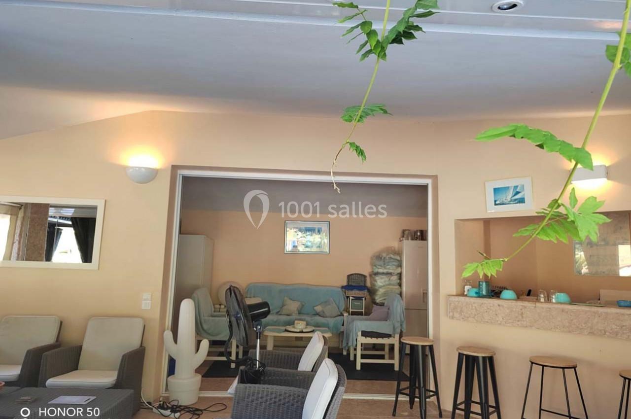 Intérieur d'une pièce lumineuse avec salon, bar, tabourets et plantes suspendues au premier plan.