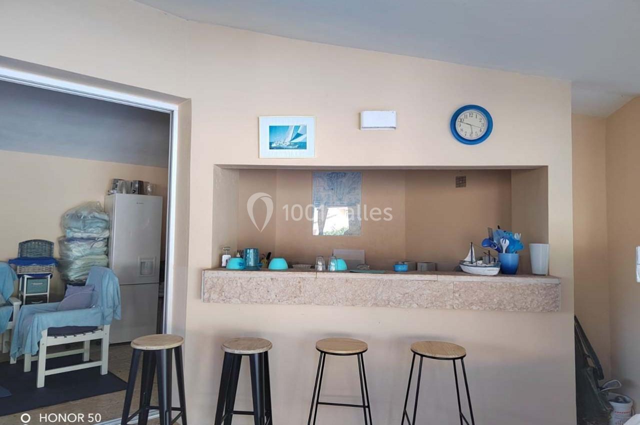 Un comptoir de cuisine avec quatre tabourets, des objets bleus et blancs, et un mur beige décoré de cadres et d'une horloge.