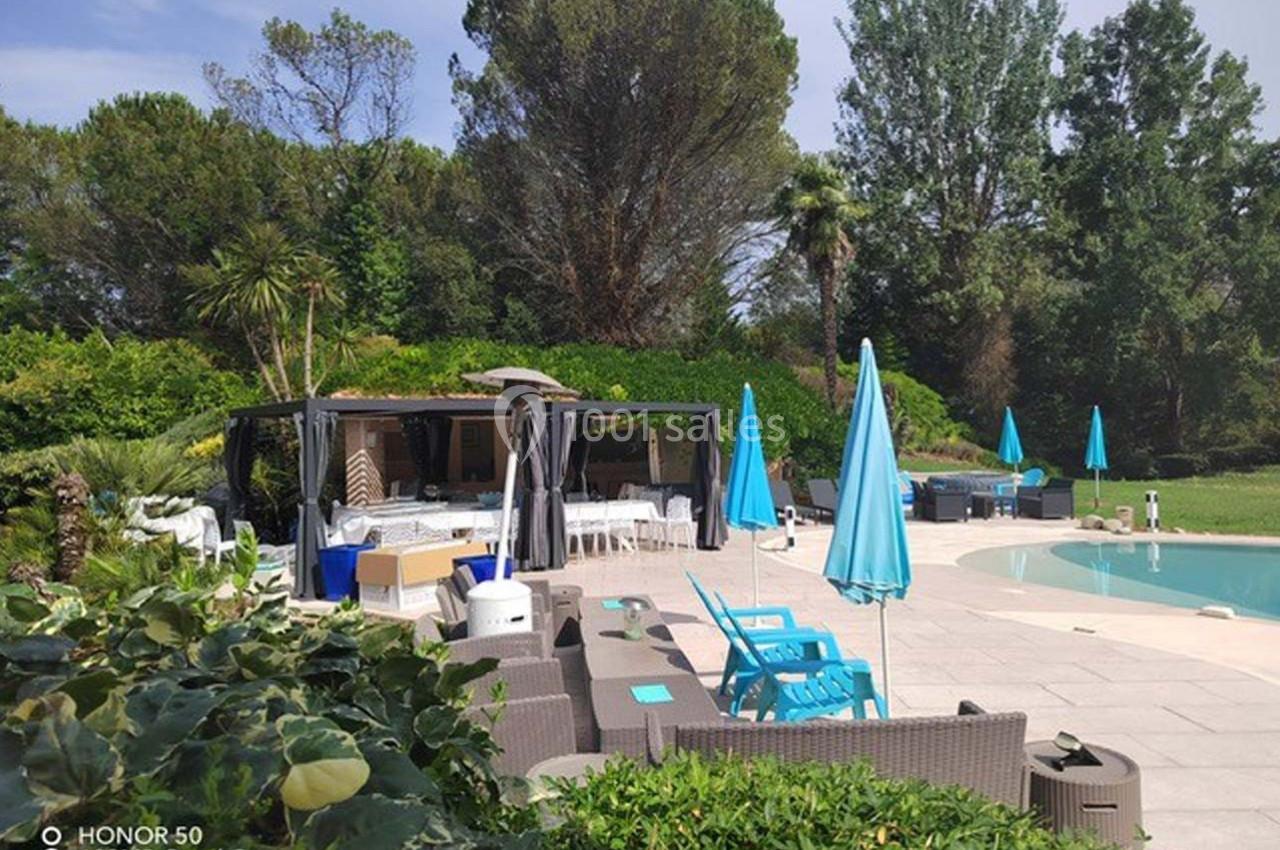 Terrasse aménagée avec mobilier de jardin, parasols bleus et piscine, entourée de végétation dense et arbres.