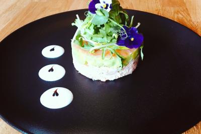 Tartare dressé avec avocat, fleurs comestibles et sauce décorative sur une assiette noire sur table en bois.