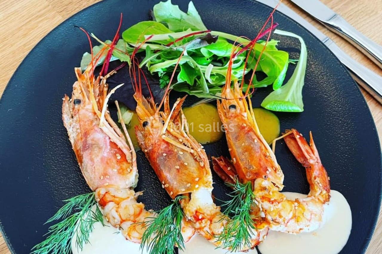 Trois gambas grillées servies avec sauce, herbes fraîches et salade verte sur une assiette noire.