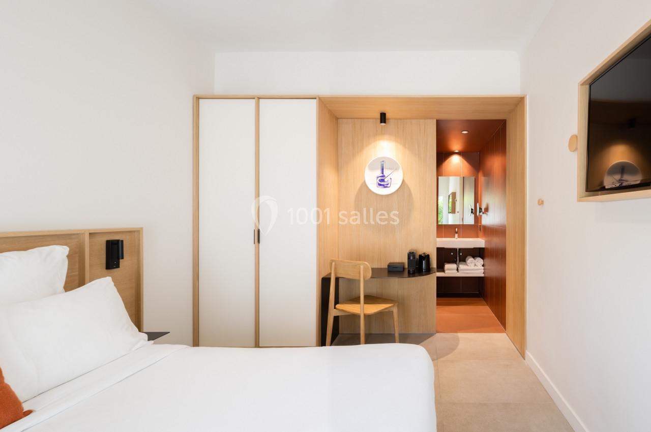 Chambre d'hôtel moderne avec lit, armoire, bureau en bois, télévision murale et salle de bain visible en arrière-plan.