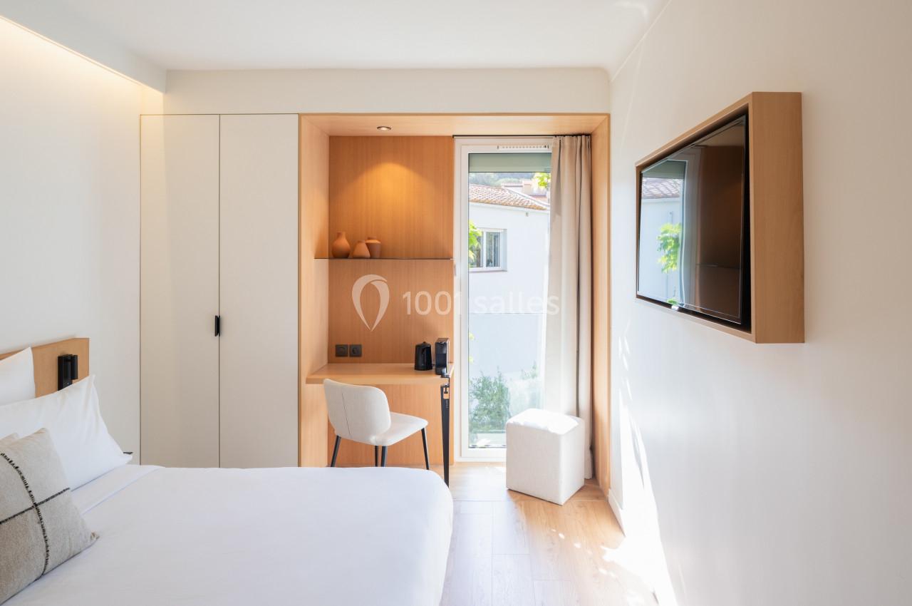 Chambre lumineuse avec lit, bureau en bois, chaise blanche, fenêtre donnant sur l'extérieur et télévision murale.