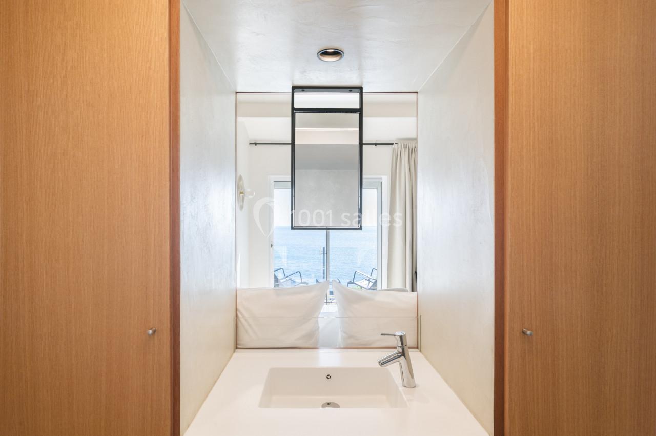 Lavabo blanc avec robinet moderne devant un miroir reflétant une vue sur la mer et des chaises sur un balcon.