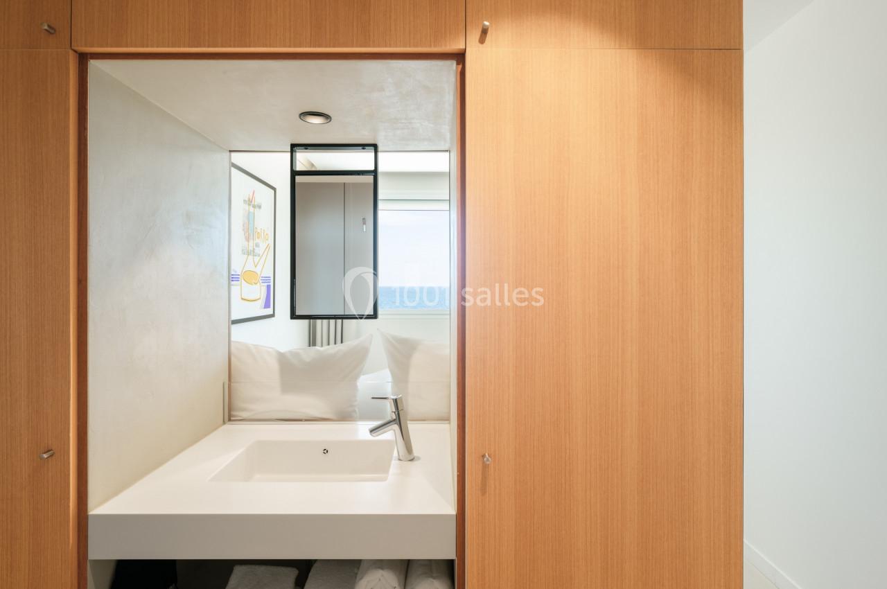 Lavabo moderne encastré dans un meuble en bois clair, avec miroir et vue sur la mer en arrière-plan.