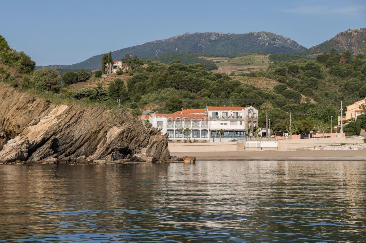 Vue d'une plage avec des bâtiments en bord de mer, des collines verdoyantes et des montagnes en arrière-plan.