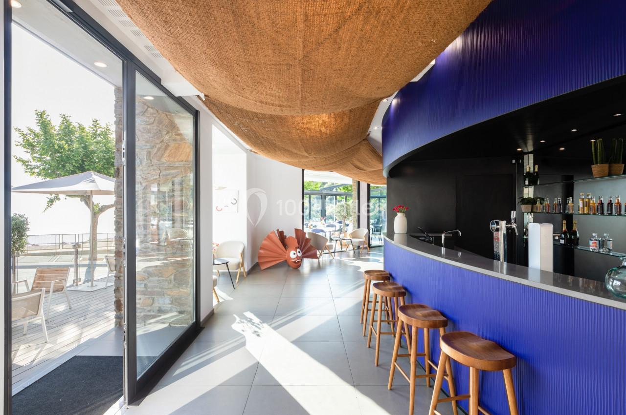 Intérieur d'un bar moderne avec comptoir bleu, tabourets en bois et grande baie vitrée donnant sur une terrasse ensoleillée.