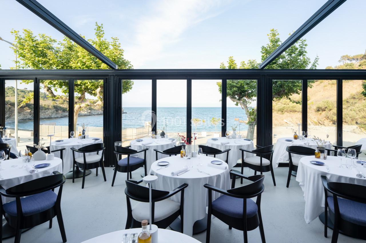 Salle de restaurant lumineuse avec baies vitrées donnant sur une vue dégagée sur la mer et des collines.