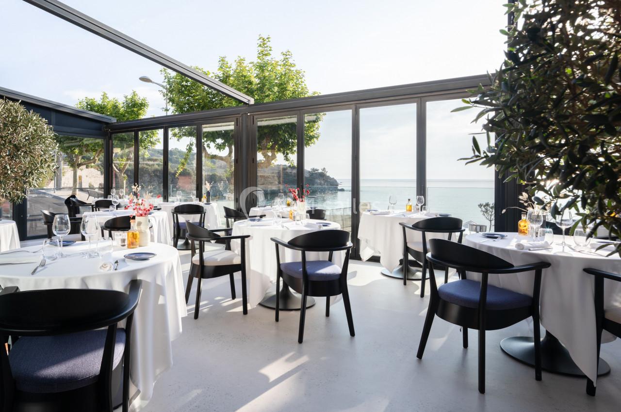 Salle de restaurant lumineuse avec baies vitrées donnant sur une vue dégagée sur la mer et des tables dressées élégamment.