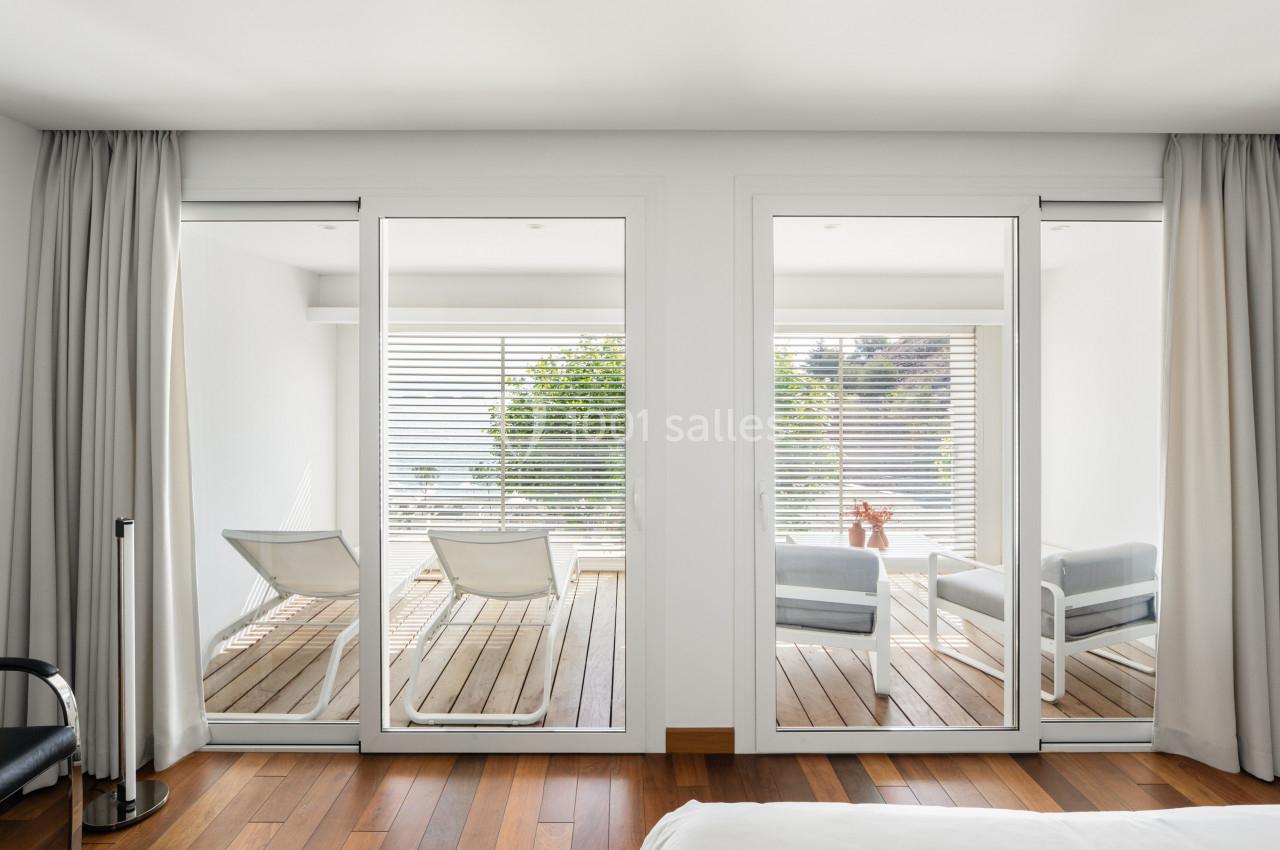 Vue depuis une chambre sur une terrasse en bois avec deux chaises longues et un fauteuil, entourée de baies vitrées.