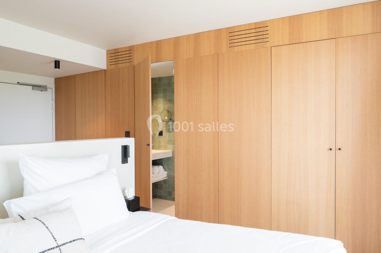 Lit avec draps blancs devant une armoire en bois, une porte ouverte révélant une salle de bain avec carrelage vert.