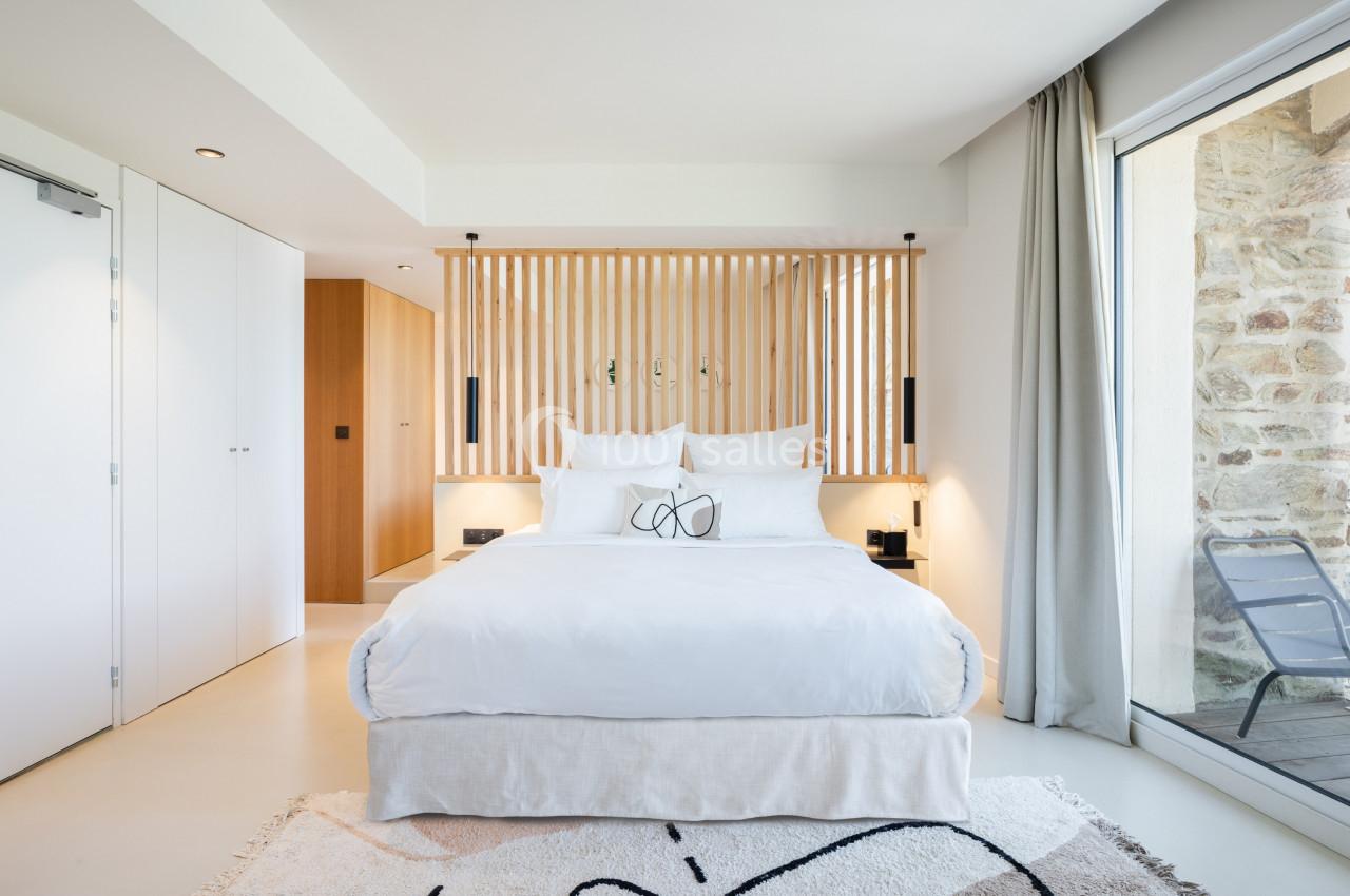 Chambre lumineuse avec lit double, tête de lit en bois ajouré, grandes fenêtres et décor minimaliste.