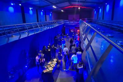 Salle lumineuse avec tables dressées pour un repas, chaises rouges, lampes suspendues et personnes en arrière-plan.