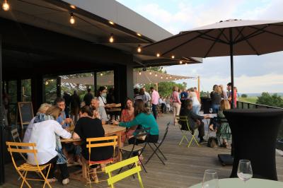 Salle lumineuse avec tables dressées pour un repas, chaises rouges, lampes suspendues et personnes en arrière-plan.