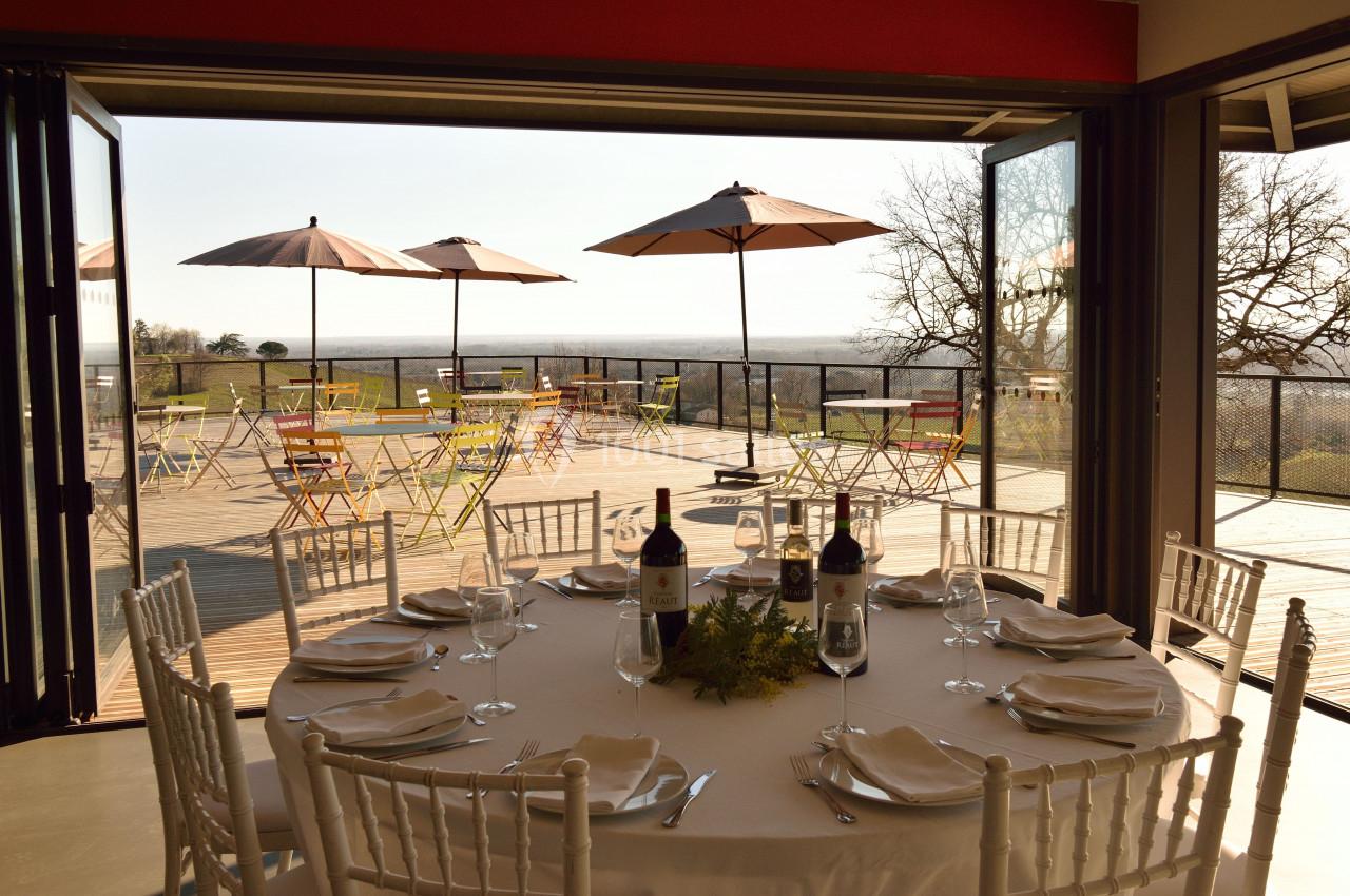 Terrasse ensoleillée avec tables et chaises colorées, vue dégagée, table dressée à l'intérieur avec bouteilles de vin.
