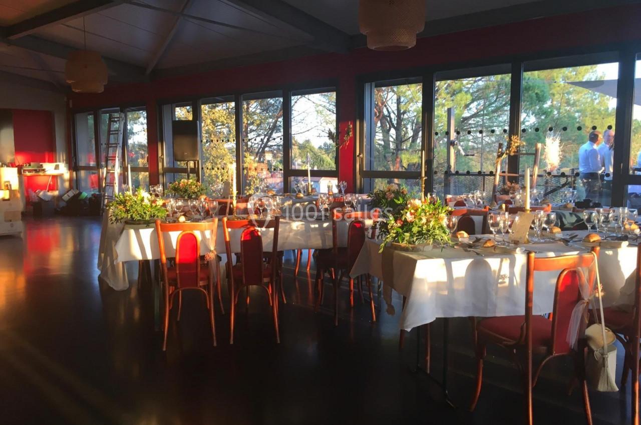 Salle de réception lumineuse avec tables décorées de fleurs et nappes blanches, entourées de chaises rouges.