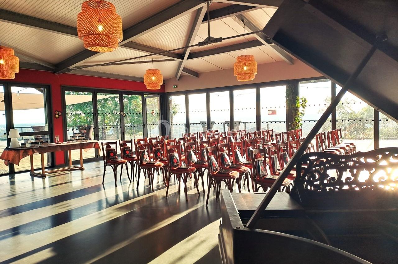 Salle lumineuse avec des rangées de chaises alignées, un piano au premier plan et des suspensions en osier au plafond.