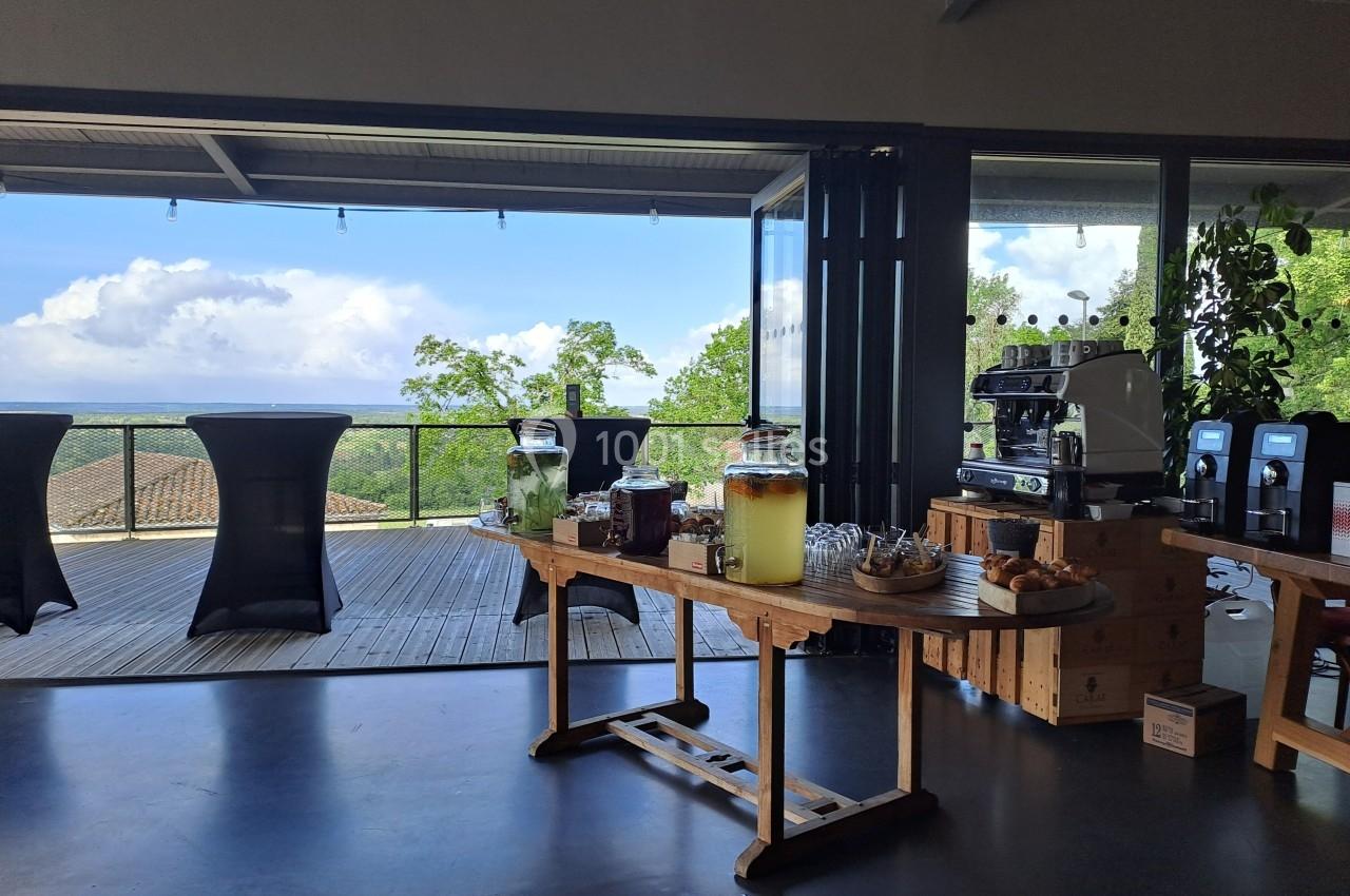 Table en bois avec boissons, viennoiseries et machine à café, située dans une salle ouverte sur une terrasse avec vue sur la…