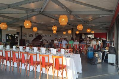 Salle lumineuse avec tables dressées pour un repas, chaises rouges, lampes suspendues et personnes en arrière-plan.