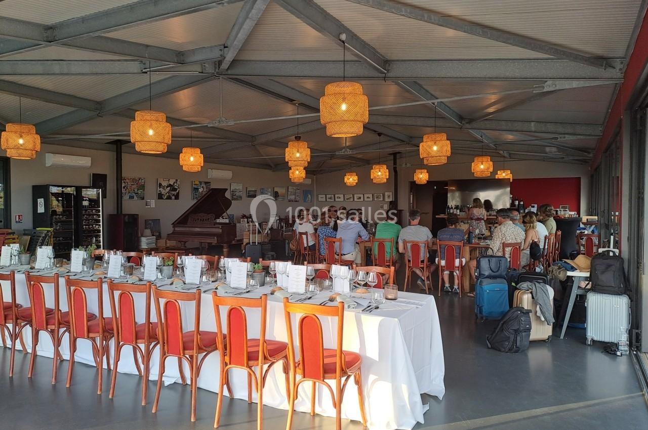 Salle lumineuse avec tables dressées pour un repas, chaises rouges, lampes suspendues et personnes en arrière-plan.
