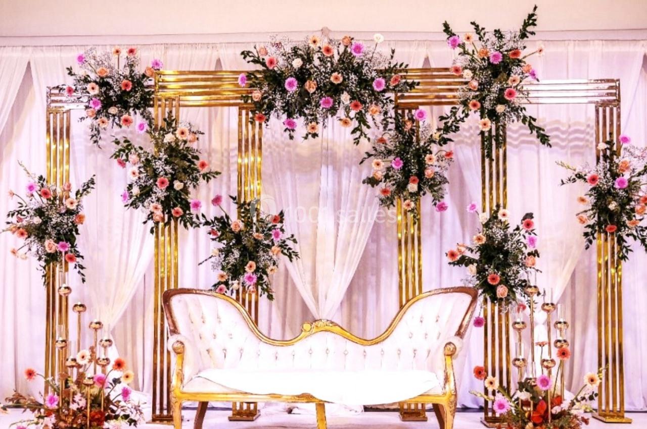 Décor de mariage avec arche dorée ornée de fleurs colorées, canapé blanc et rideaux blancs en arrière-plan.