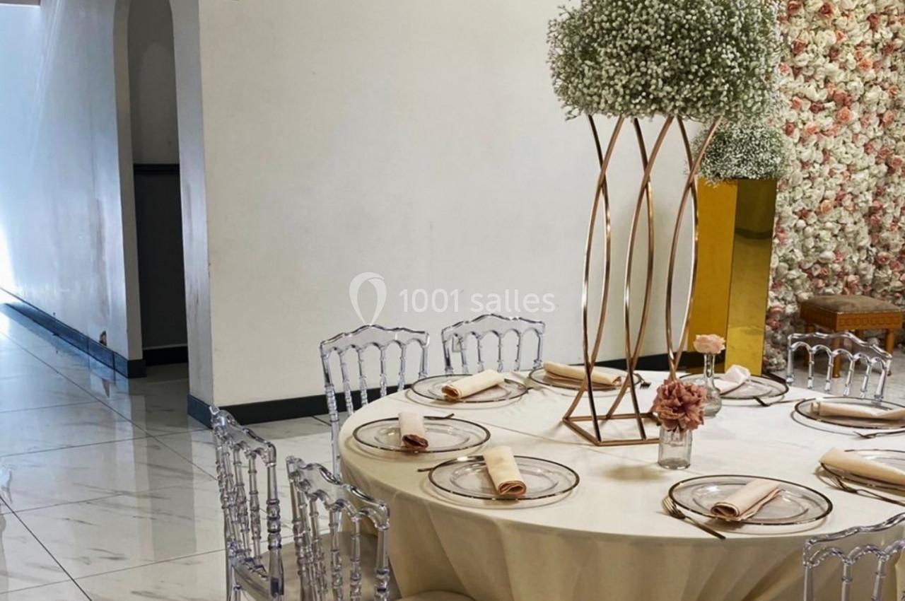 Salle de réception élégante avec table dressée, chaises transparentes et décoration florale sur fond de mur fleuri.