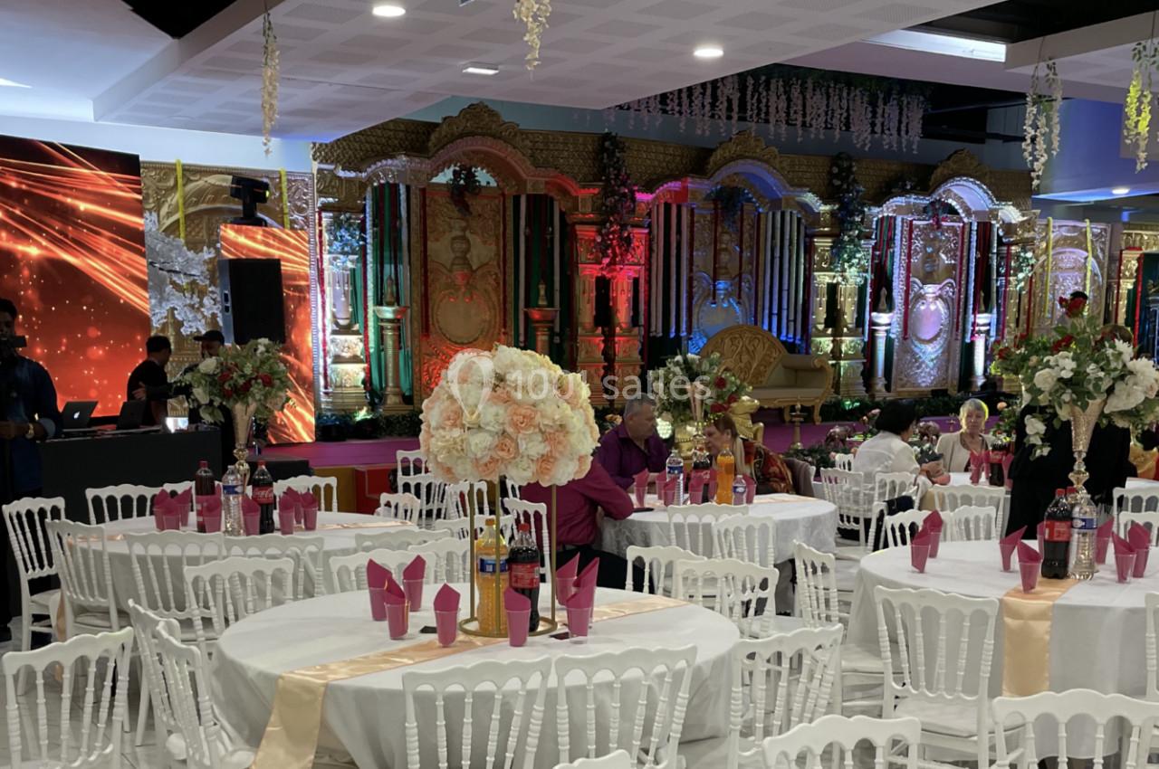 Salle de réception décorée avec des tables blanches, des fleurs et une scène ornée de lumières colorées en arrière-plan.