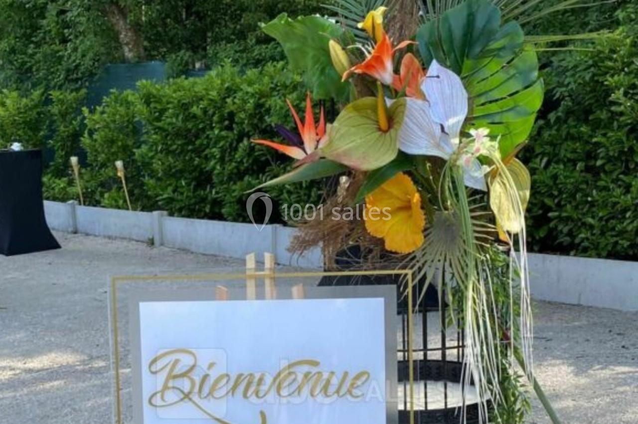 Panneau ’Bienvenue’ décoré de fleurs tropicales colorées, placé à l'extérieur près d'une haie et de tables hautes noires.