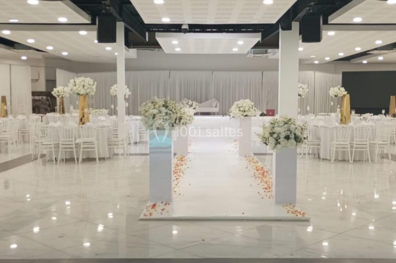 Salle de réception élégante avec tables rondes dressées, allée centrale décorée de fleurs blanches et pétales au sol.
