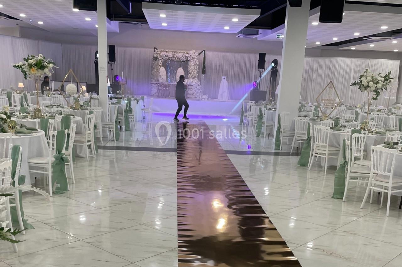 Salle de réception décorée avec des tables rondes, des chaises blanches et une allée centrale menant à une scène ornée.