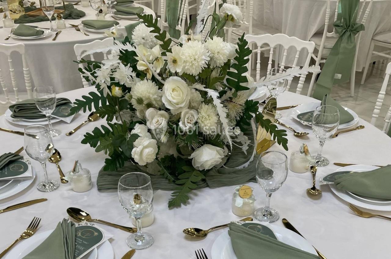 Table ronde élégamment dressée avec une nappe blanche, serviettes vertes, couverts dorés et un centre de table floral blanc.