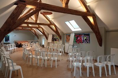 Salle lumineuse avec poutres apparentes, tables rondes dressées avec nappes blanches et chaises en plastique blanc.