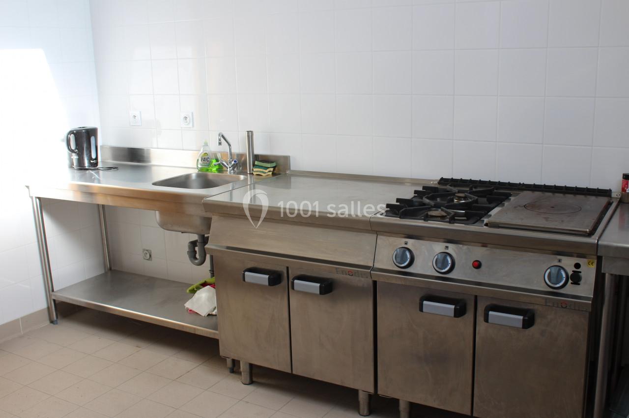 Cuisine professionnelle avec plan de travail en inox, évier, bouilloire, plaques de cuisson et carrelage mural blanc.